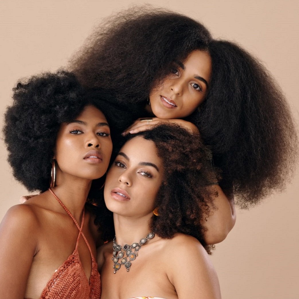 Three women with diverse skin tones and beautiful, voluminous curly hair styles stand together.